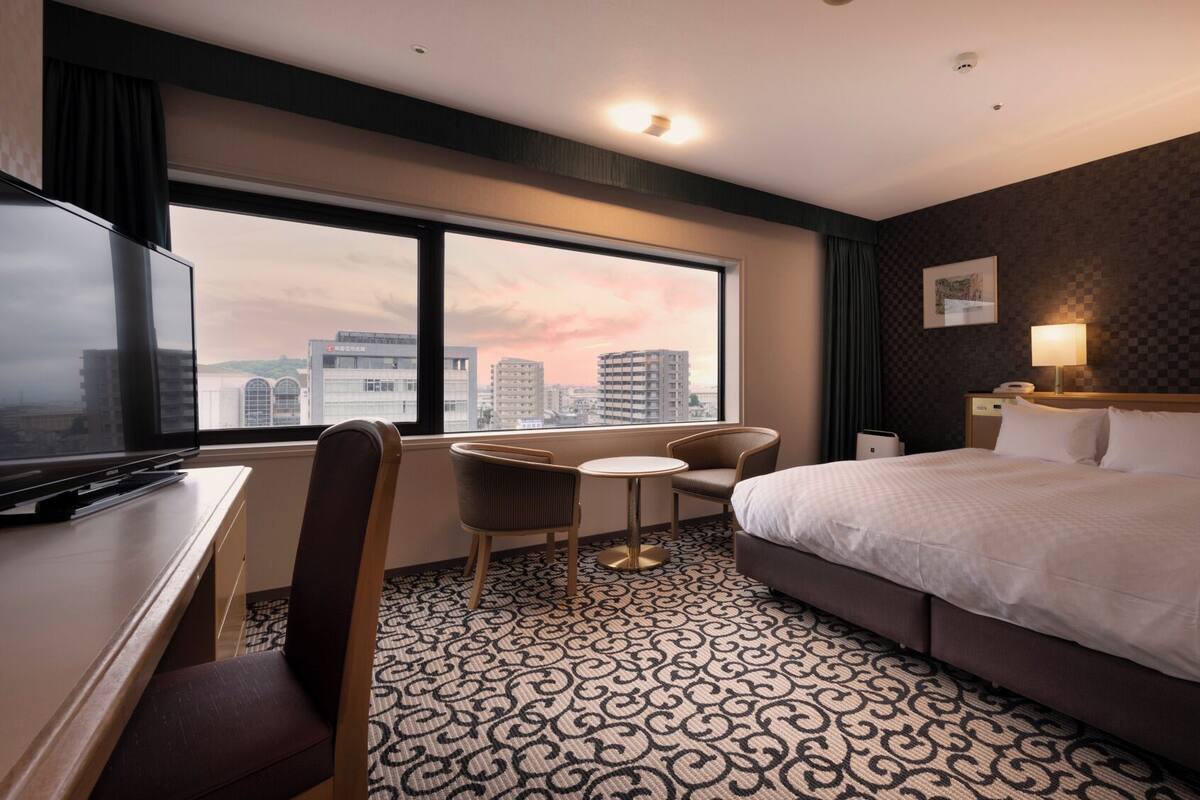 Ein Hotelzimmer mit einem großen Bett, einem Schreibtisch mit Fernseher, einem Sessel, einem kleinen Tisch und Blick auf die Stadt durch große Fenster.