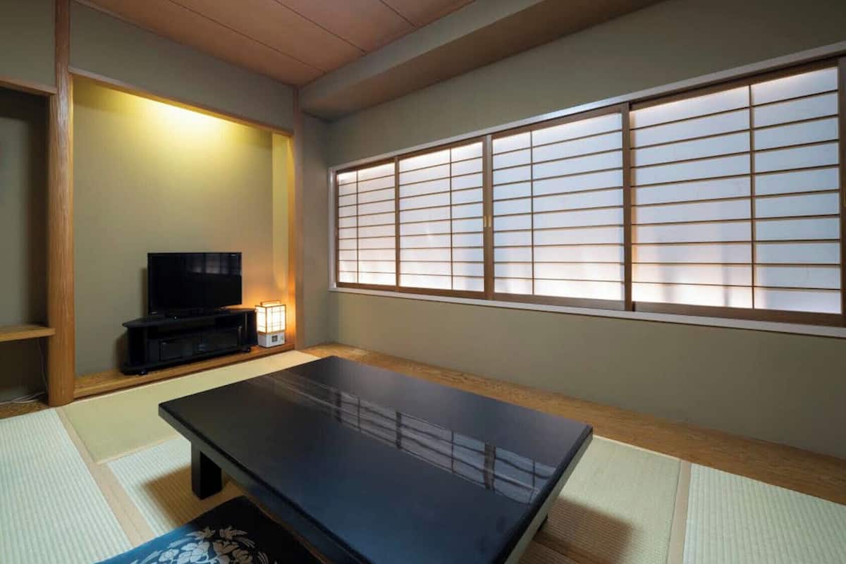 Una habitación japonesa tradicional con suelo de tatami, una mesa negra, un televisor y un celosía japonesa.