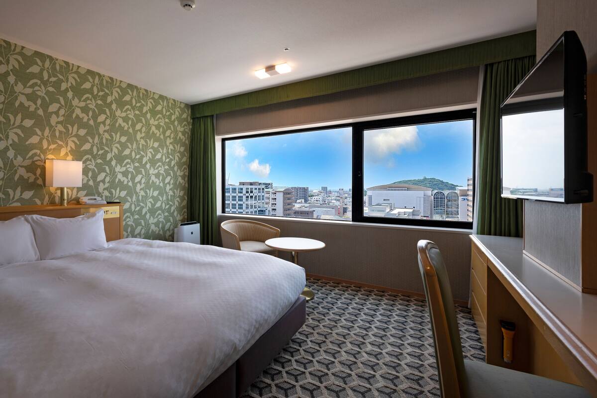 Ein Hotelzimmer mit Bett, Schreibtisch, Stuhl, einem kleinen Tisch und einem Fenster mit Blick auf die Stadt.