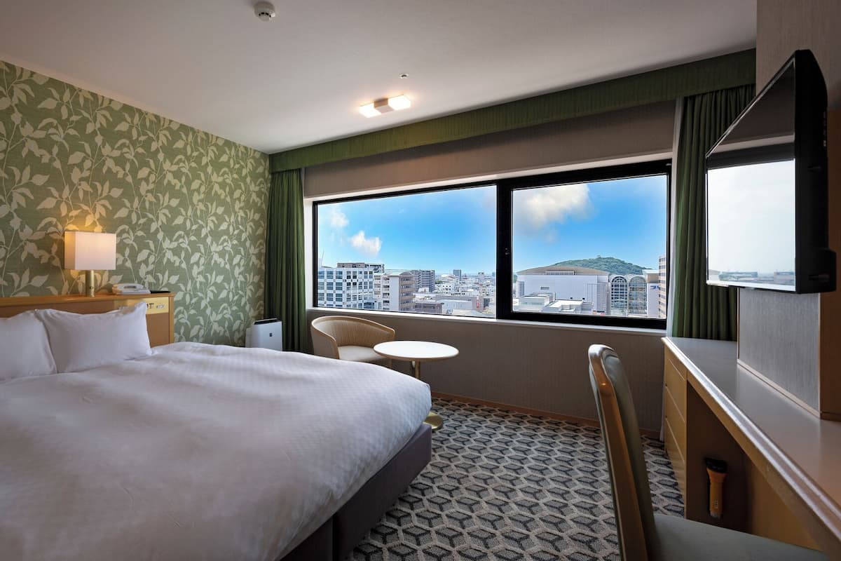 Ein Hotelzimmer mit einem Bett, einem Sessel, einem kleinen Tisch, einer Lampe, einem Fenster mit Stadtblick und einer Deckenleuchte.