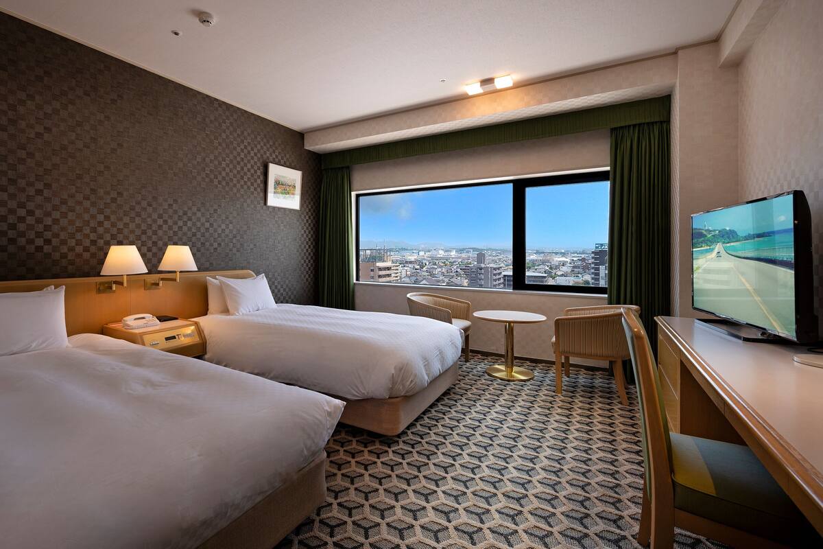 Ein Hotelzimmer mit zwei Betten, einem Schreibtisch, einem Stuhl, einem Fernseher und einem großen Fenster mit Blick auf die Stadt.