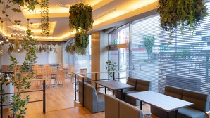 Interior moderno de restaurante com mesas e cadeiras de madeira, janelas amplas com persianas e plantas penduradas.