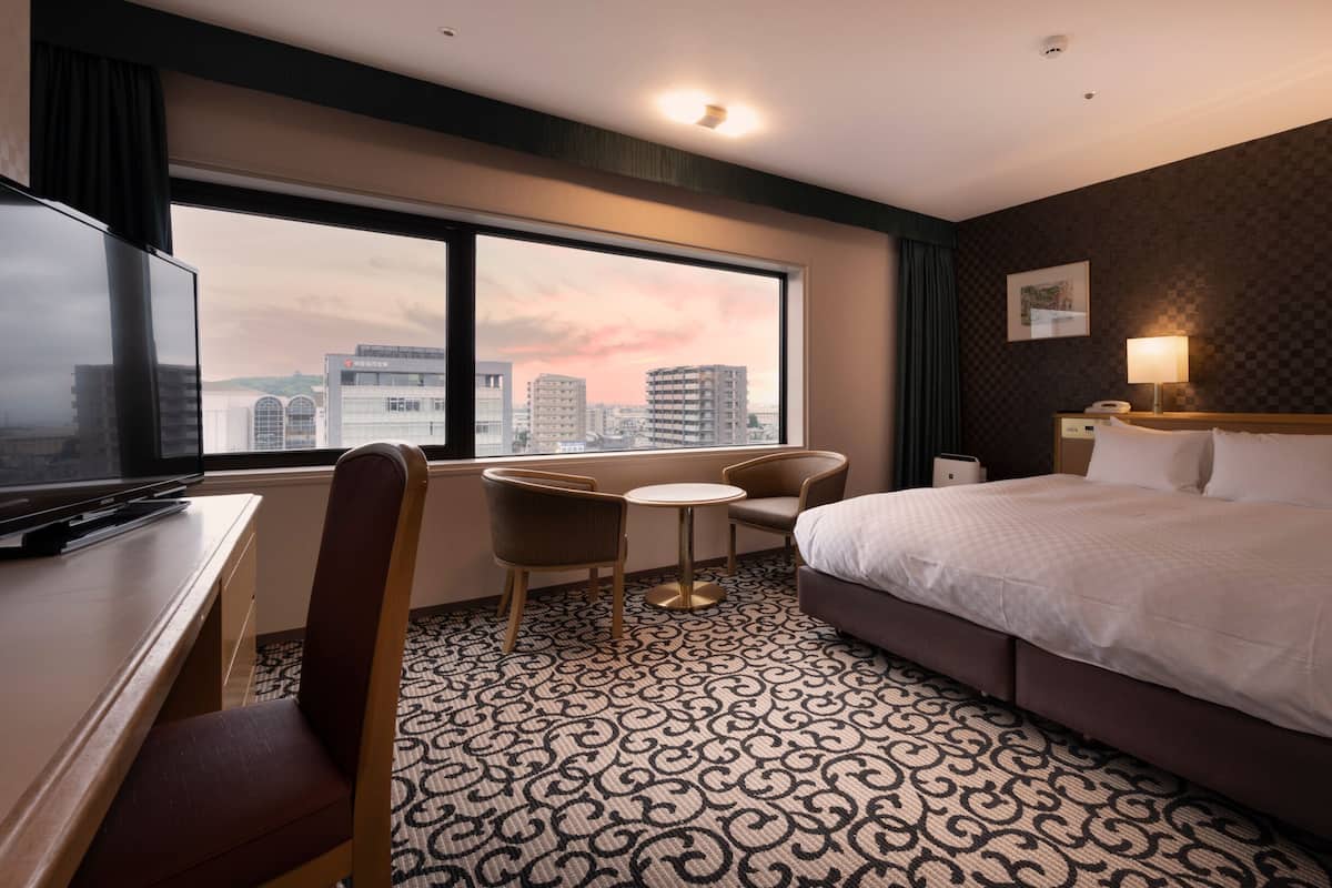 Ein Hotelzimmer mit einem großen Bett, einem Schreibtisch mit Fernseher, einem Sessel, einem kleinen Tisch und Blick auf die Stadt.