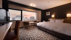 Quarto de hotel com uma cama grande, uma escrivaninha com televisão, uma cadeira, uma mesinha e vista para a cidade pela janela.