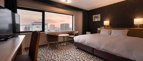 A hotel room with a large bed, a desk with a TV, a chair, a small table, and a view of the cityscape through the window.