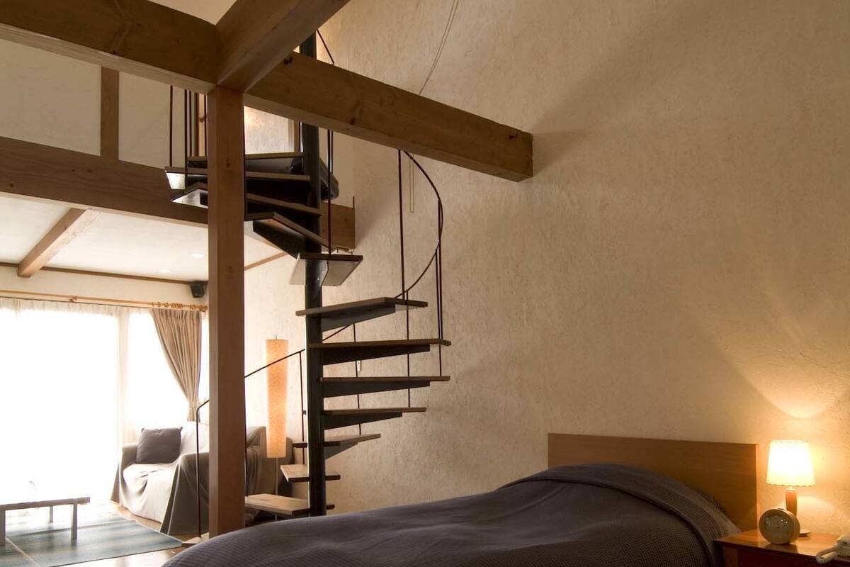A bedroom with two beds, wooden beams, a spiral staircase, a bedside table with a lamp, and a view of a balcony.