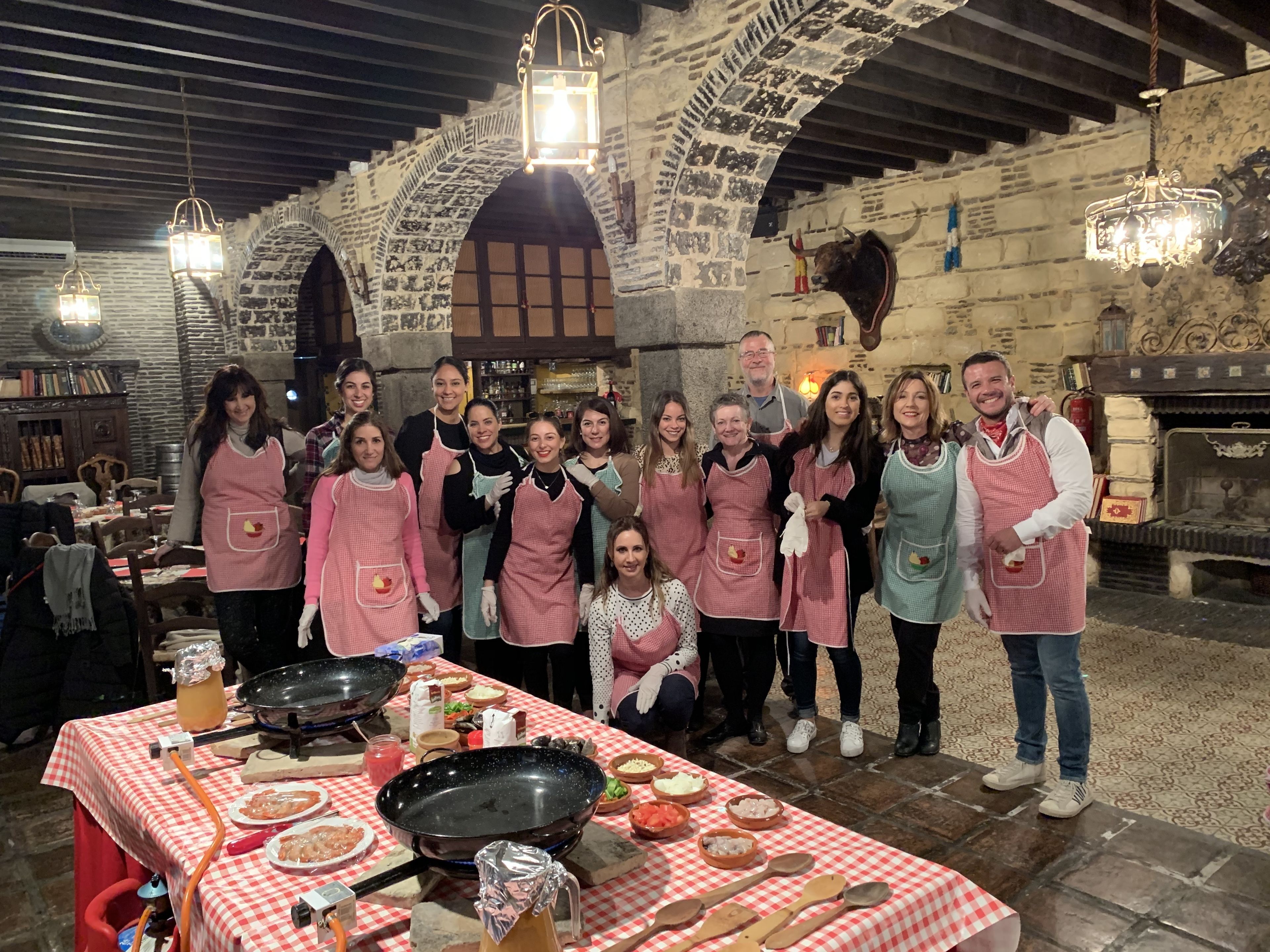 A group of people in aprons gathered around a table with cooking utensils and food items in a rustic stone-walled kitchen.