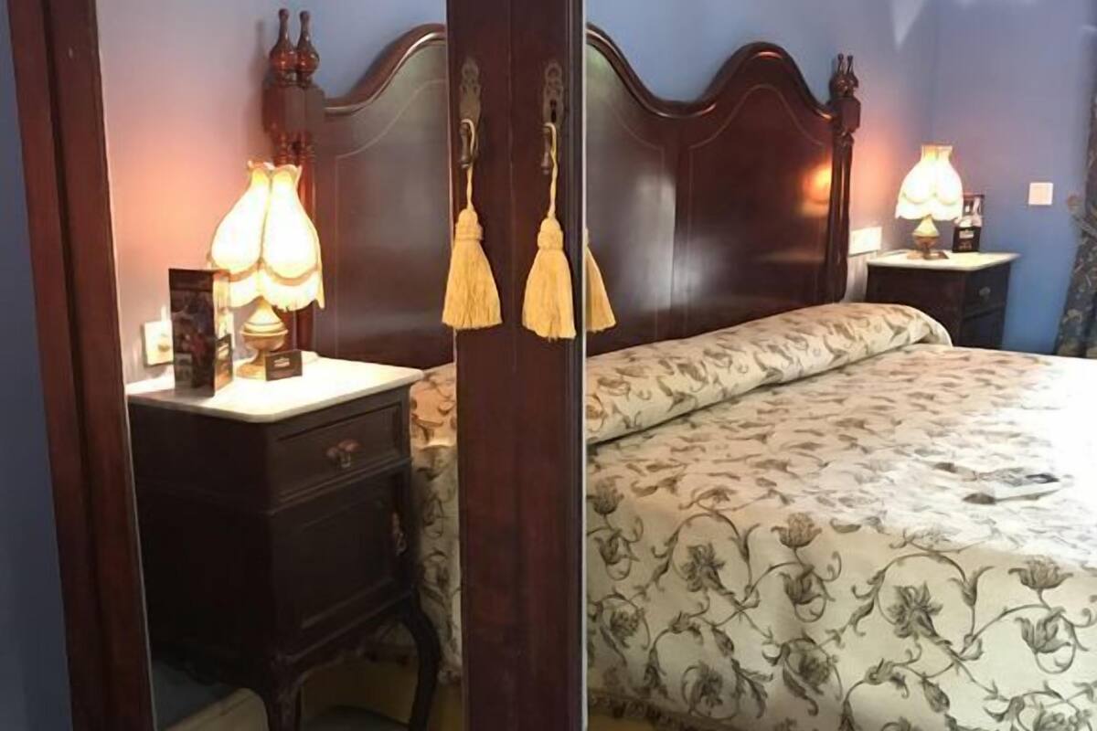 A bedroom with a large mirror reflecting a bed with floral bedding, a nightstand with a lamp, and a dresser with a mirror.