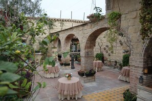 Un patio al aire libre con arcos de piedra, mesas redondas con manteles rosa y plantas en macetas.