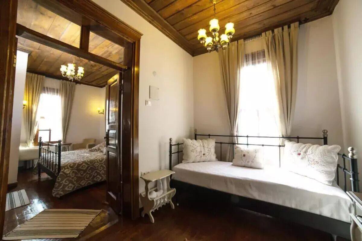 A bedroom with a large bed, wooden ceiling beams, a chandelier, and a view of another room.
