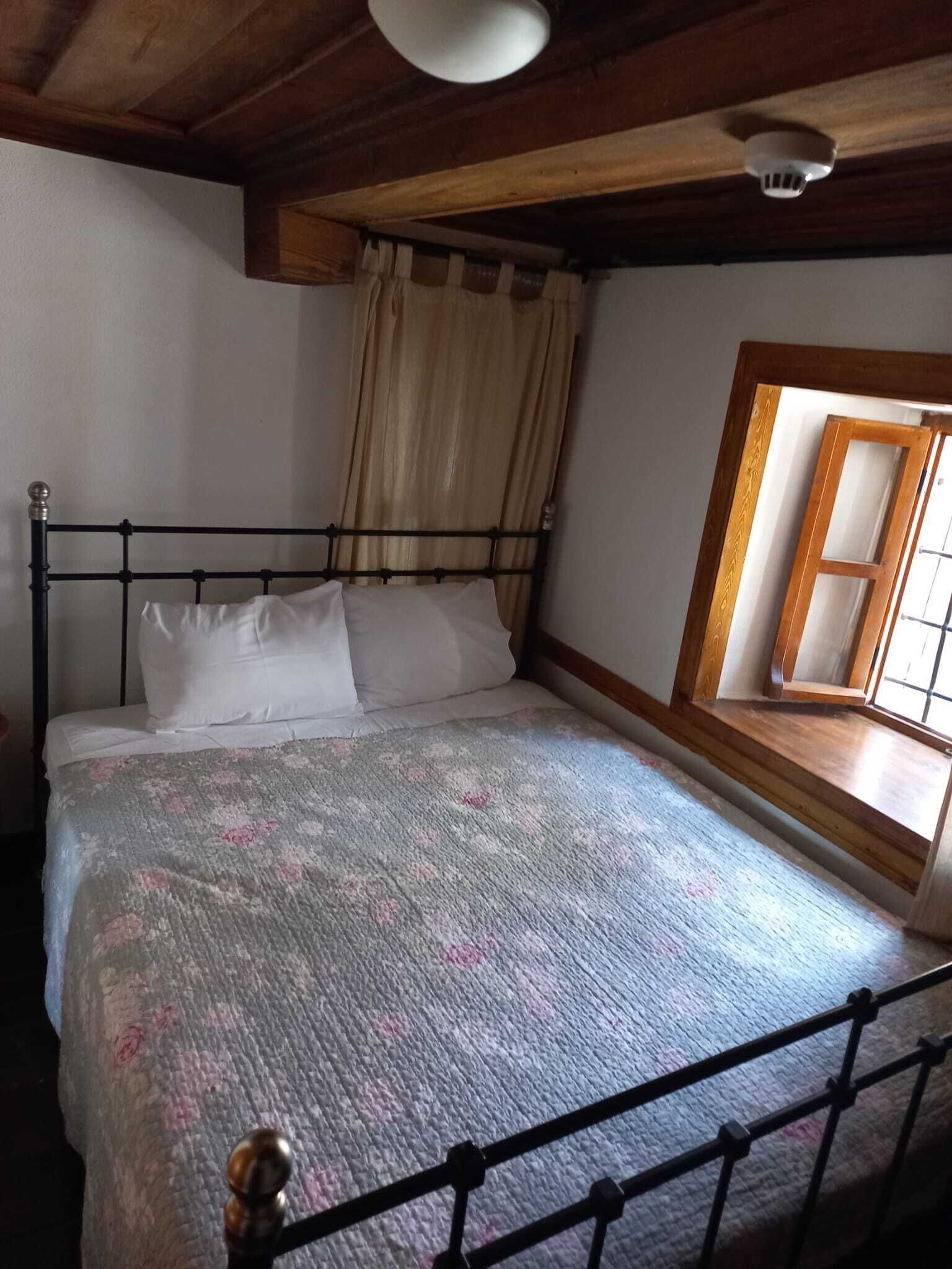 A bedroom with a metal bed frame, a floral bedspread, a window with a wooden frame, and a ceiling fan.