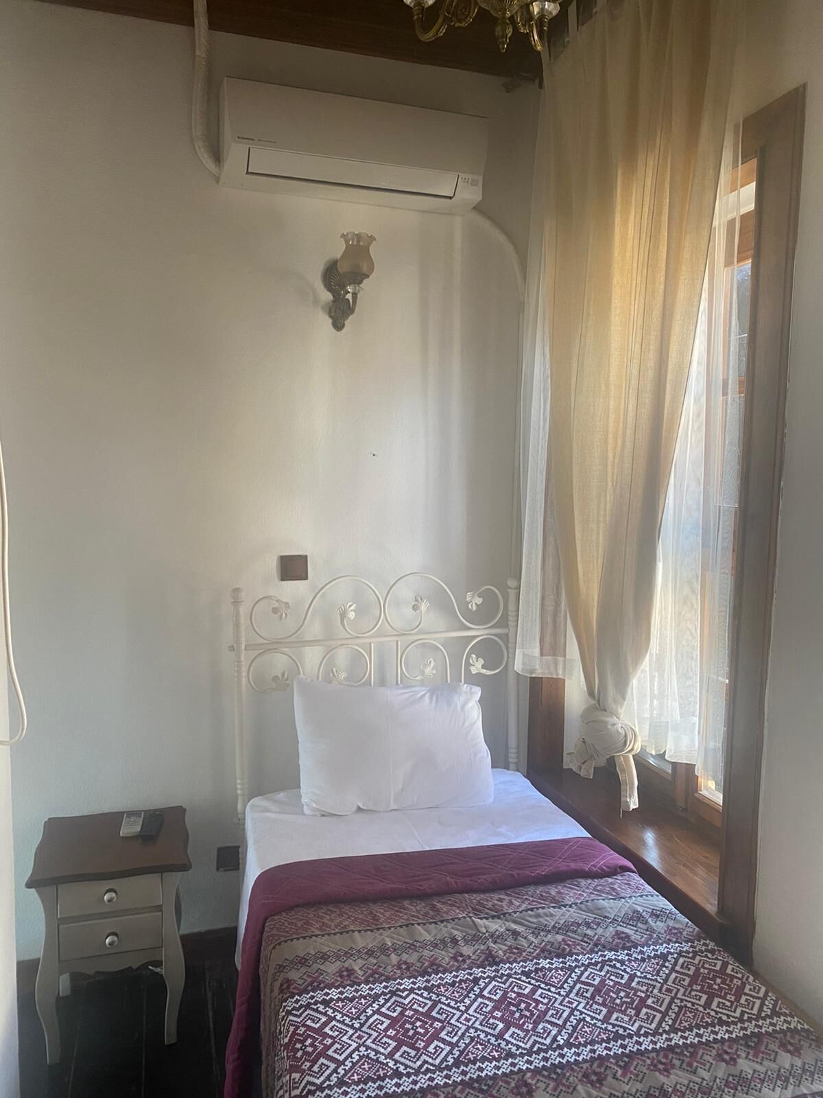 A bedroom with a bed, a nightstand, a wall-mounted air conditioner, and a window with sheer curtains.