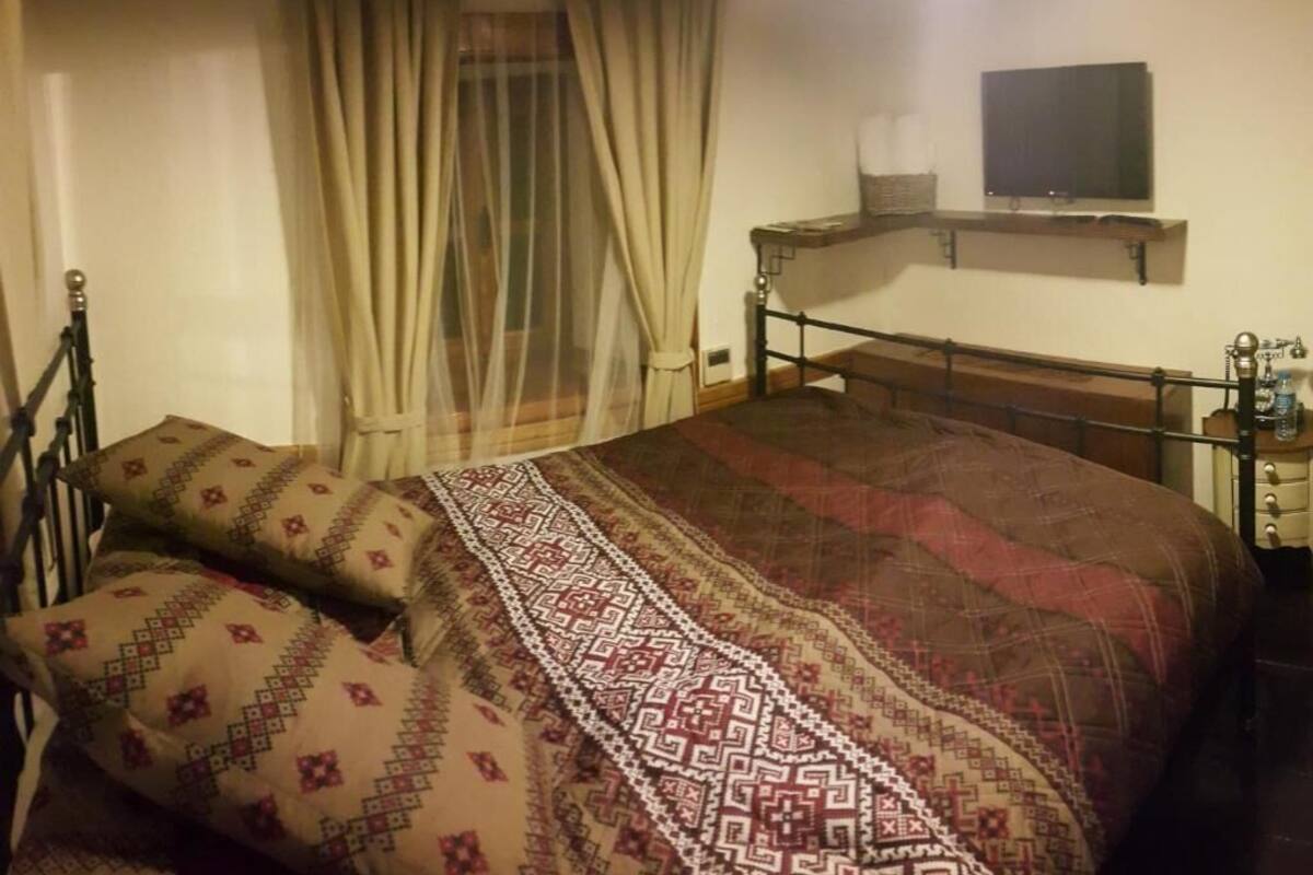 A bedroom with a bed, a bedside table, a TV, and a window with curtains.