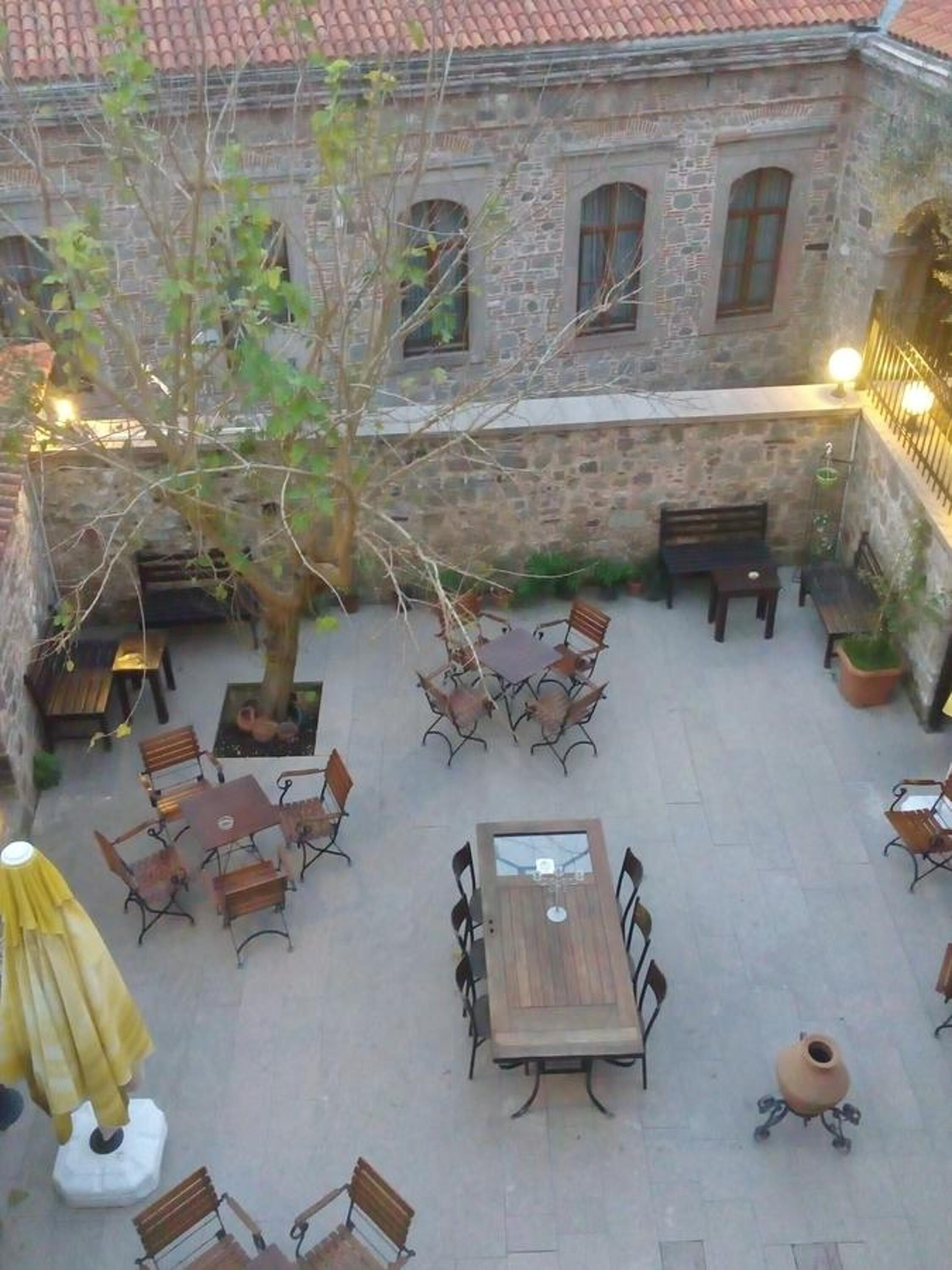 An outdoor patio area with stone walls, multiple tables and chairs, and a tree providing shade.