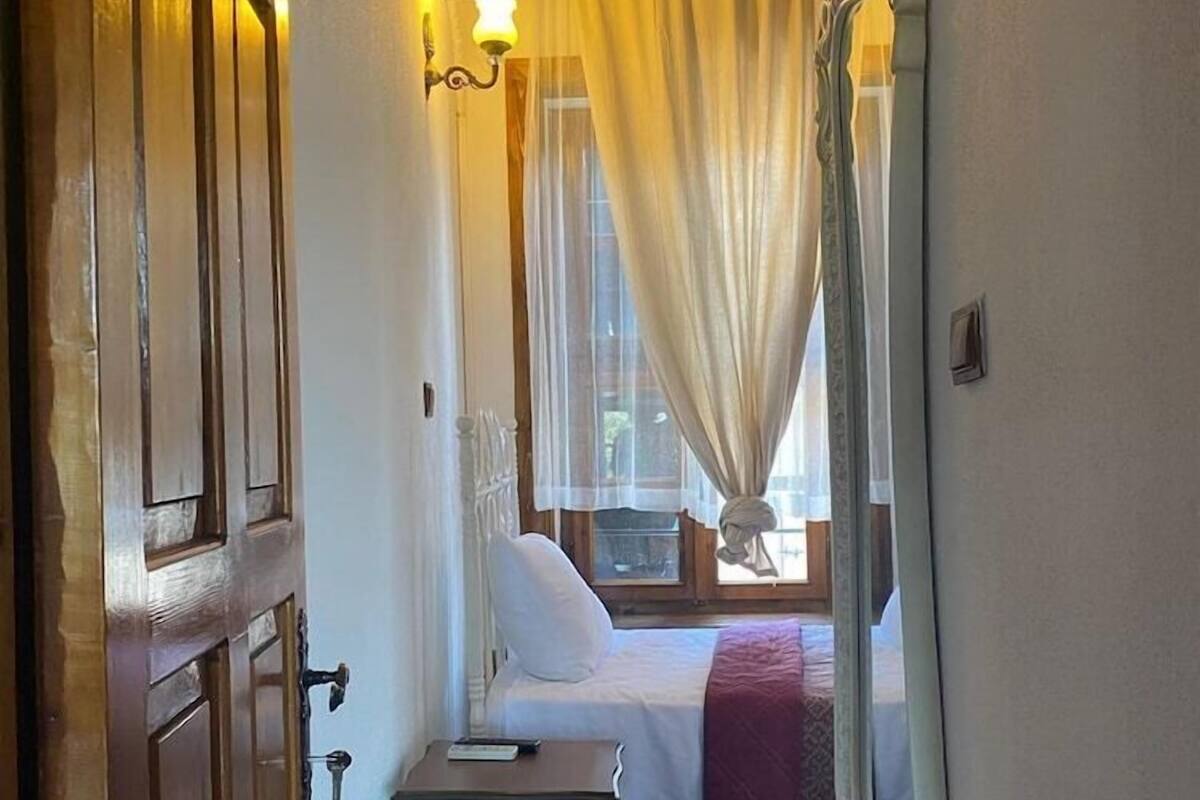 A narrow hallway leading to a bedroom with a bed, a bedside table, a mirror, and a window with curtains.