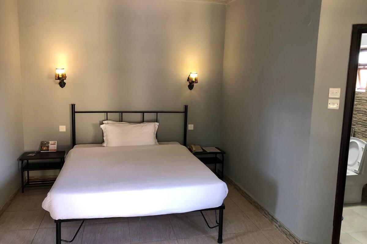 A single bed with a white mattress and black metal frame, flanked by two bedside tables with lamps, in a room with light grey walls and a tiled floor.