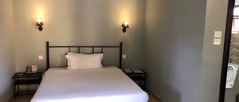 A single bed with a white mattress and black metal frame, flanked by two bedside tables with lamps, in a room with light grey walls and a tiled floor.