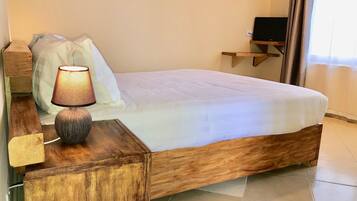 Een net opgemaakt bed met een houten hoofdbord, een bedlampje en een televisie op een dressoir tegen de muur.