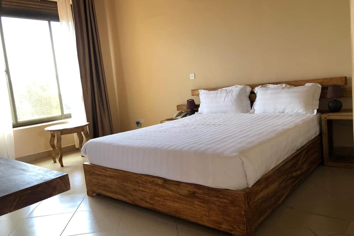 A large bed with a wooden frame and white bedding, a small bedside table, a lamp, and a window with curtains.