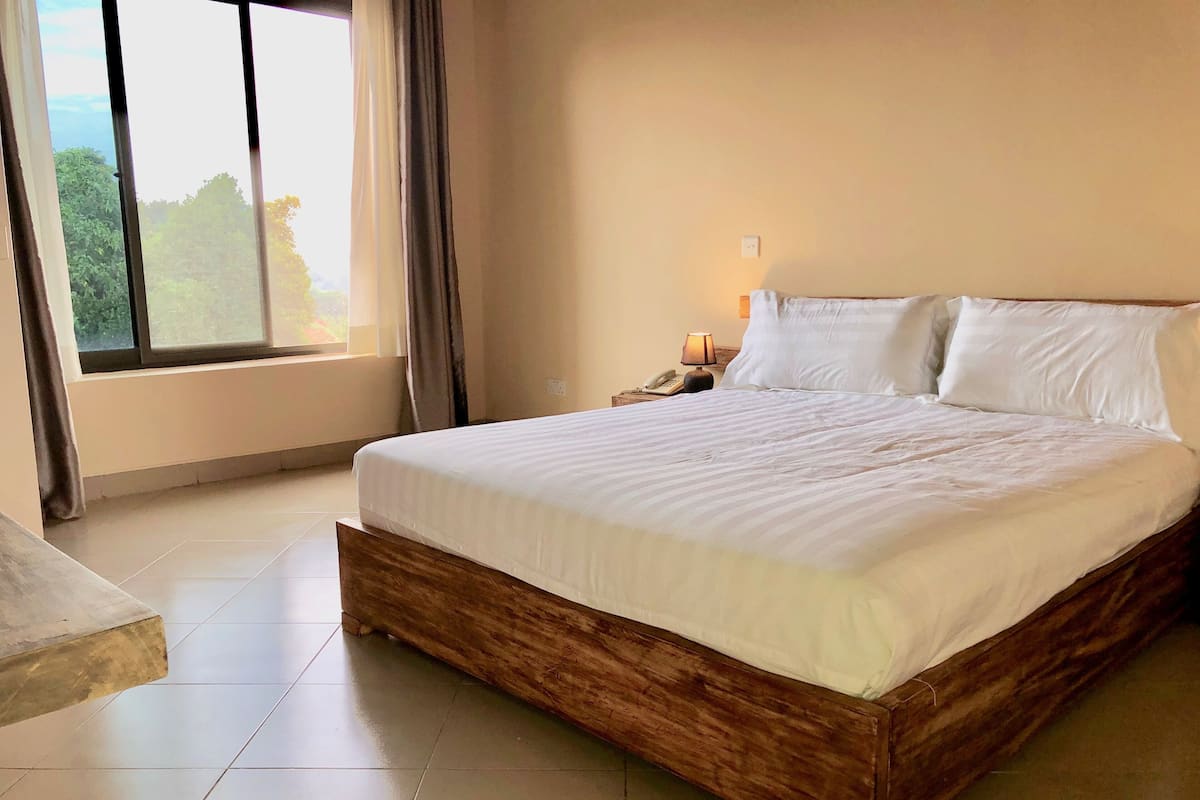 A neatly made bed with white linens in a room with a large window, a bedside table with a lamp, and a telephone.