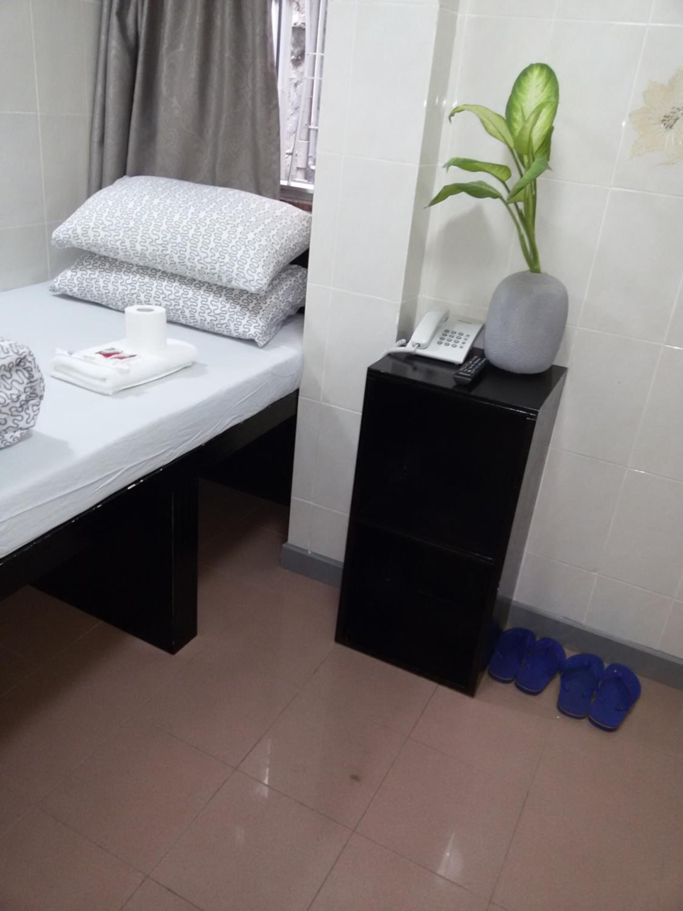 Een kleine, verzorgde kamer met een bed, een nachtkastje met een telefoon en een plant, en een paar slippers op de vloer.