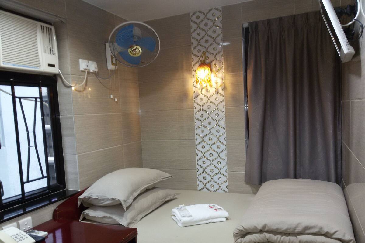 A hotel room with a bed, a desk with a telephone, a wall-mounted fan, and a window with blinds.