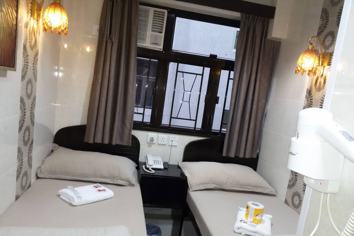 A hotel room with two beds, a window with curtains, a bedside table with a phone, and a hairdryer.