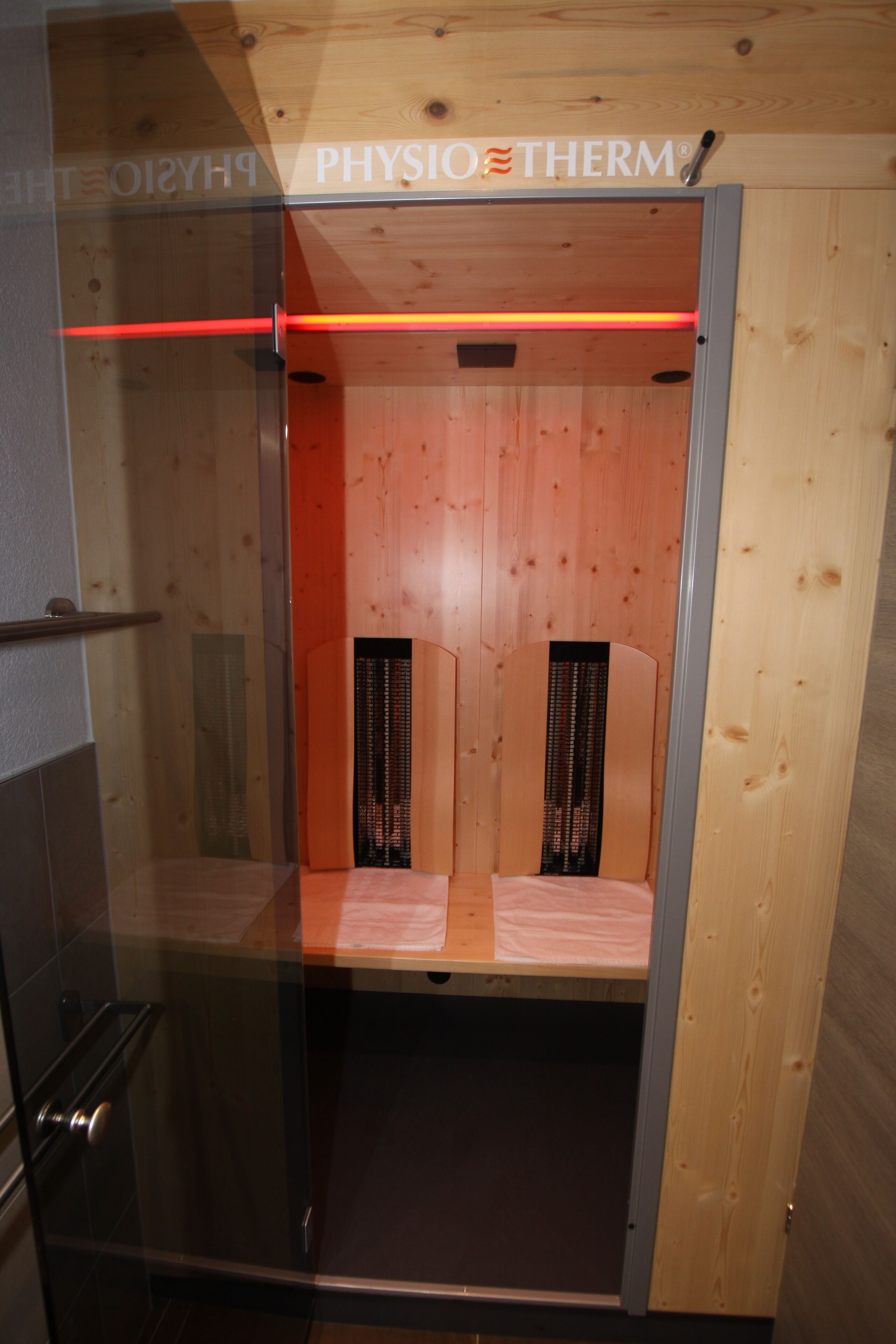 Een sauna met houten wanden en een glazen deur, met een rode lichtbalk en een zichtbaar merk.