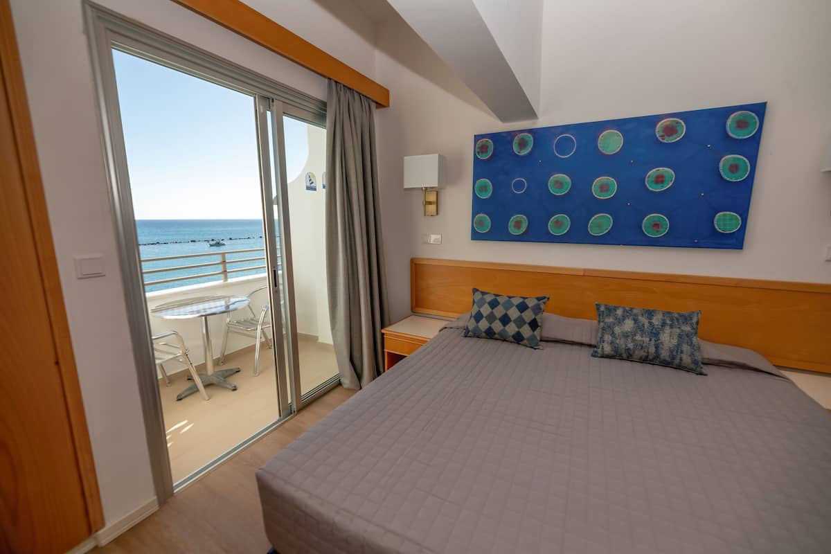 A hotel room with a bed, a nightstand, a lamp, a large abstract painting, and a view of the sea through a sliding door.