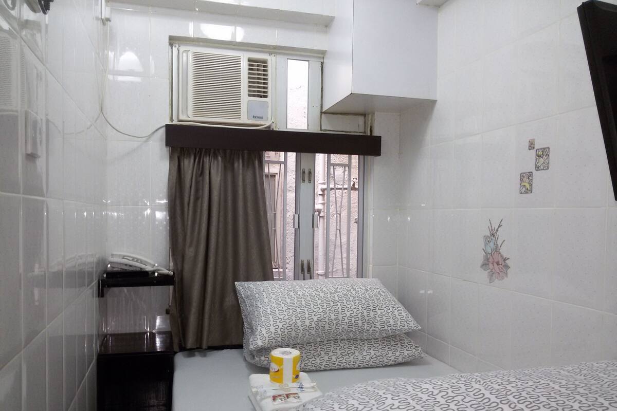 A small, clean room with a bed, a window with curtains, a radiator, and a wall with a small decorative item.