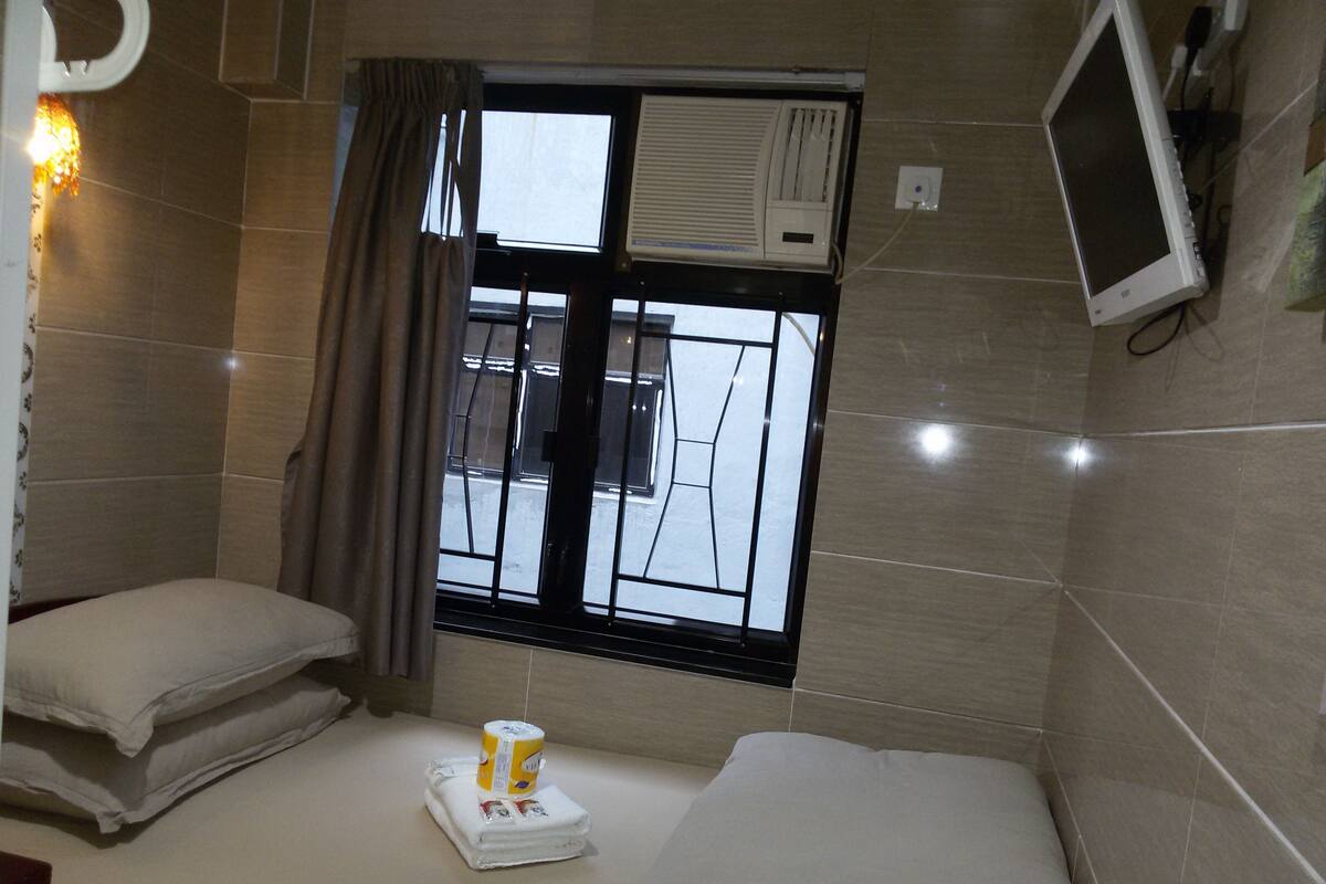 A hotel room with a bed, a window with curtains, an air conditioning unit, and a television mounted on the wall.