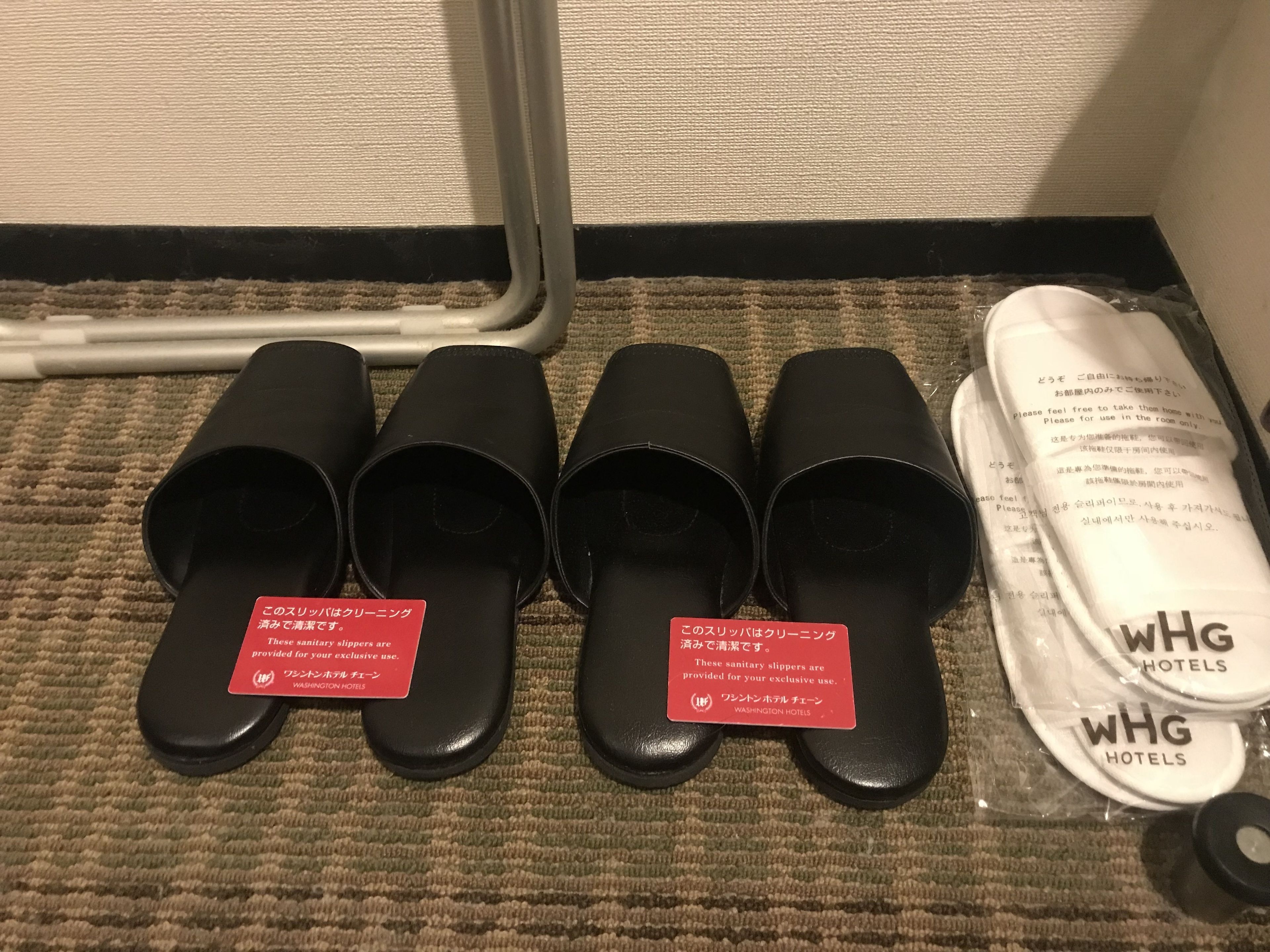 A set of black slippers with red tags on a carpeted floor, next to a WHG Hotels branded item.