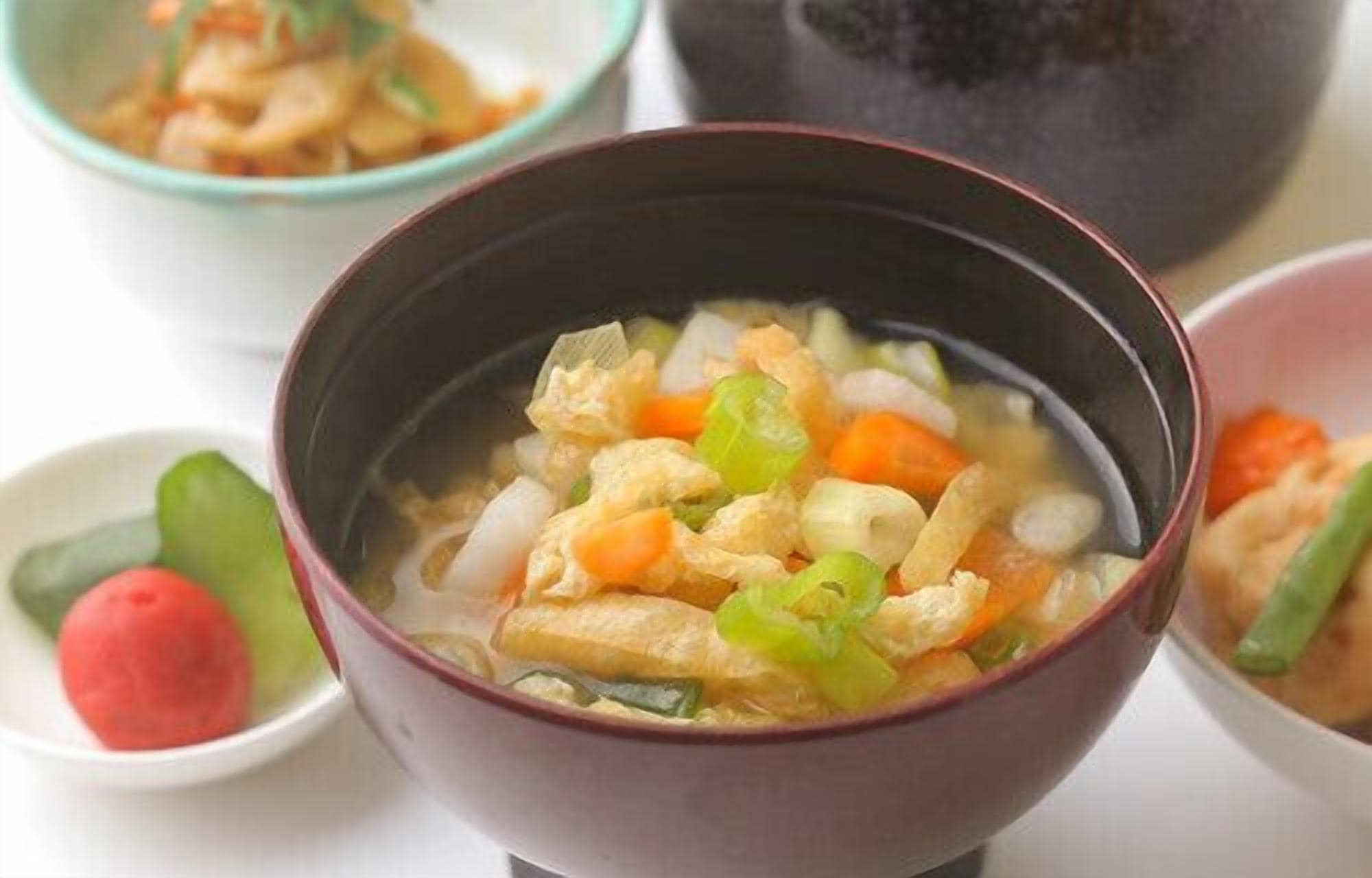 A bowl of soup with vegetables and tofu, accompanied by a small dish of pickled vegetables and a red fruit.