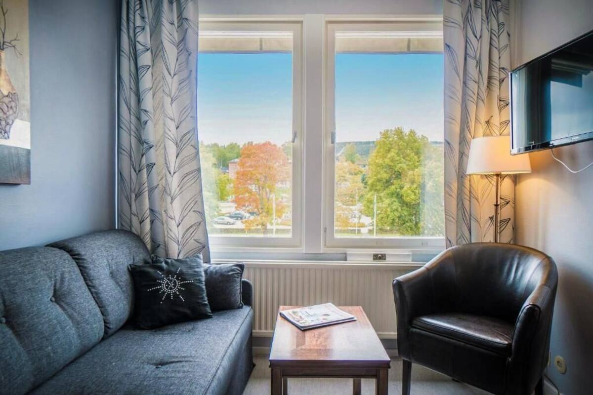 A living room with a grey sofa, a black leather chair, a wooden coffee table, and a window with a view of trees and buildings.