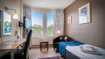 Una habitación de hotel con cama, escritorio con silla, televisor, ventana con cortinas e imagen enmarcada en la pared.