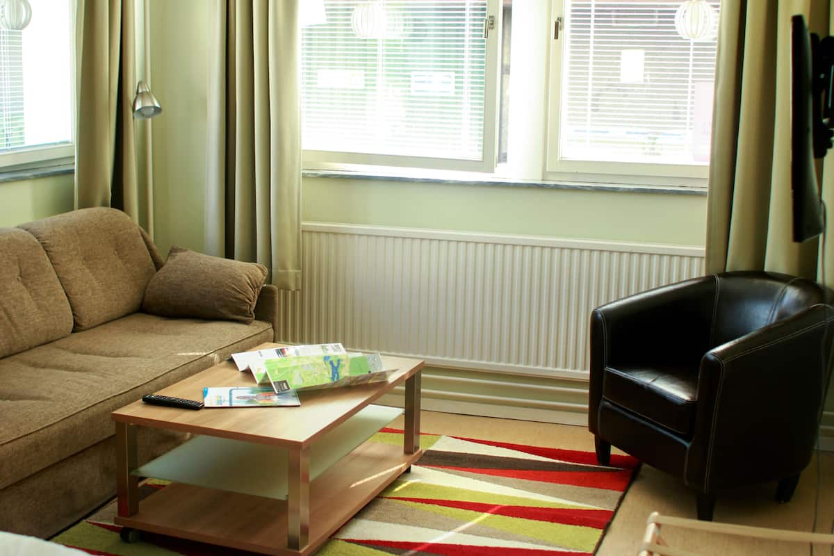 A living room with a sofa, a coffee table with magazines, a black leather chair, and a window with blinds.