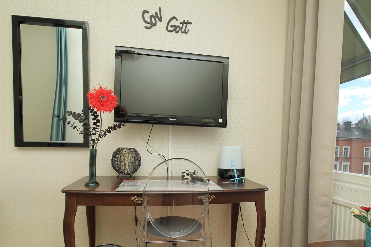 A room with a mounted TV, a desk with a vase of flowers, and a window with a view of buildings.