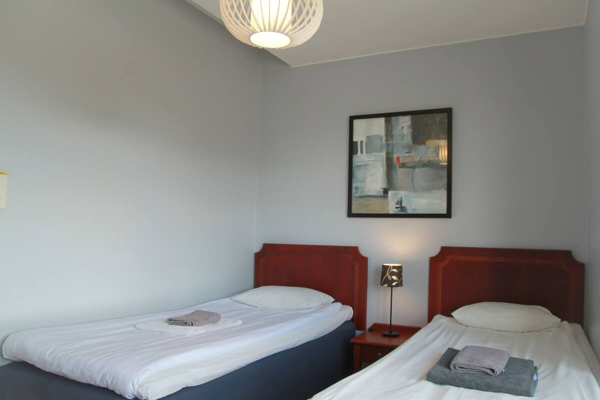 A hotel room with two single beds, a nightstand with a lamp, and a painting on the wall.