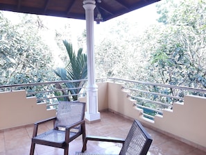 A covered balcony with two chairs, a white column, and a view of dense foliage. - Vinses Villa in Hikkaduwa (Gonapinuwala)