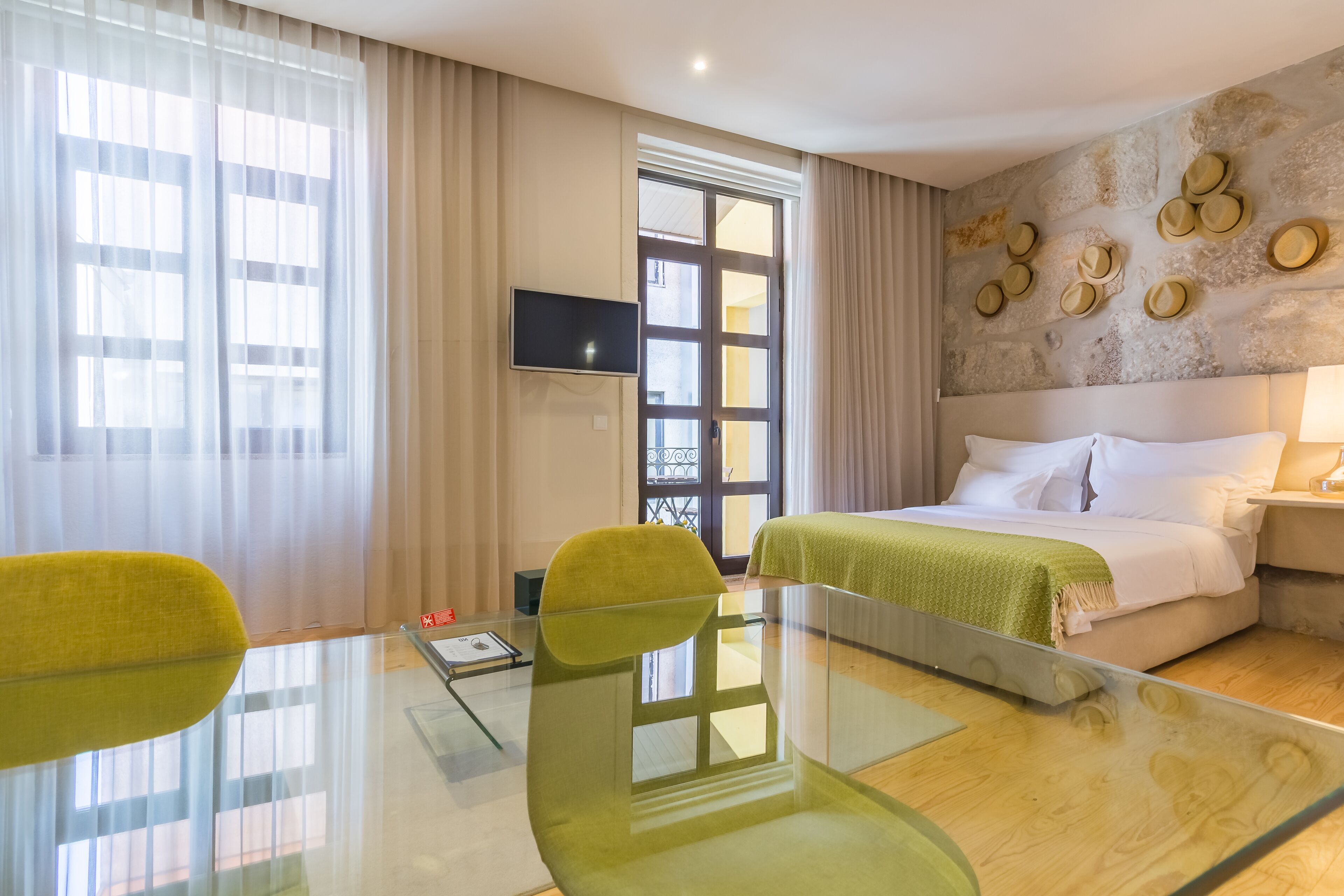 A hotel room with a glass table, two green chairs, a bed with white linens, a TV on the wall, and a stone wall with decorative plates.