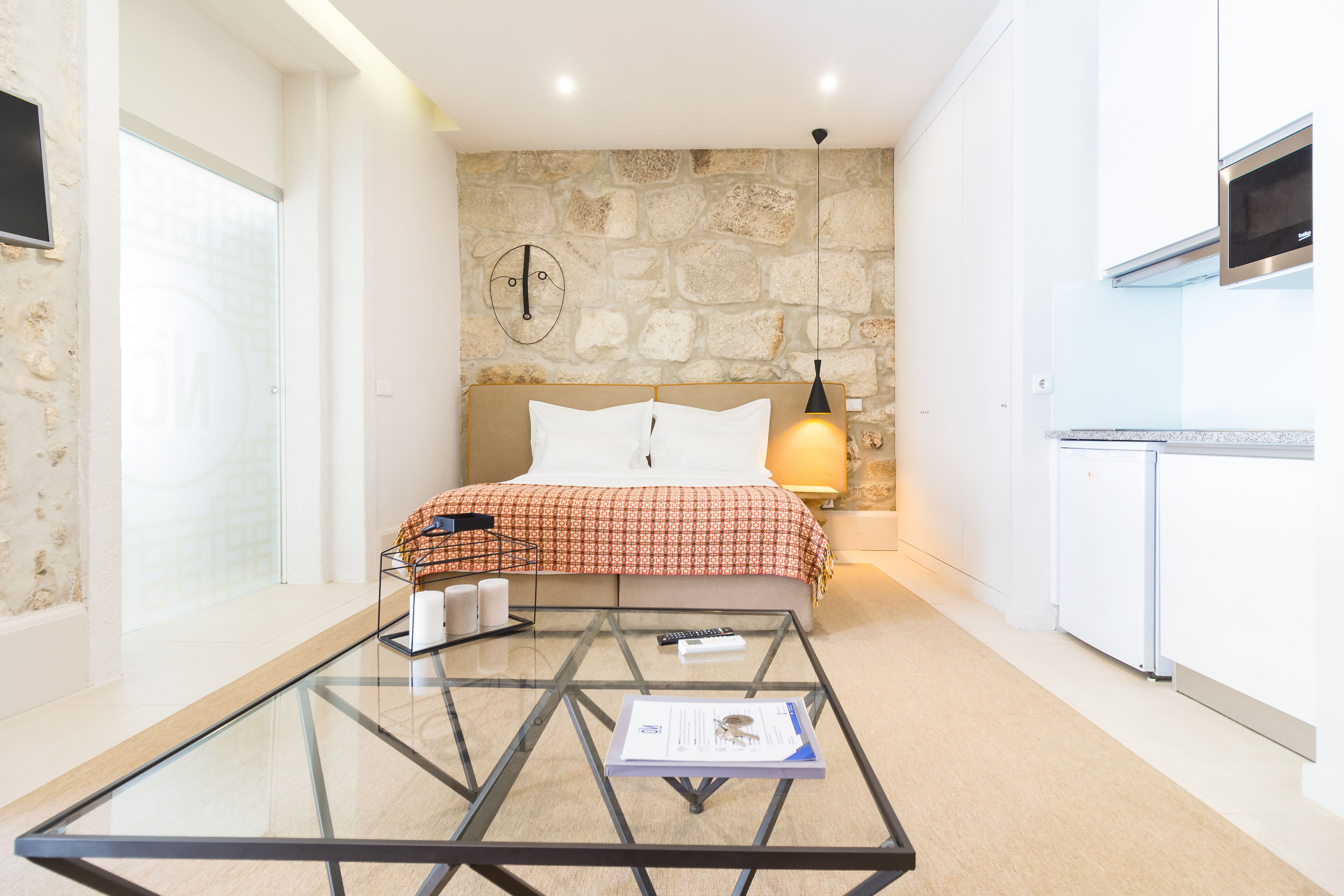 A bedroom with a glass coffee table, a bed with a red and white checkered bedspread, a stone wall, and a kitchenette with a microwave.