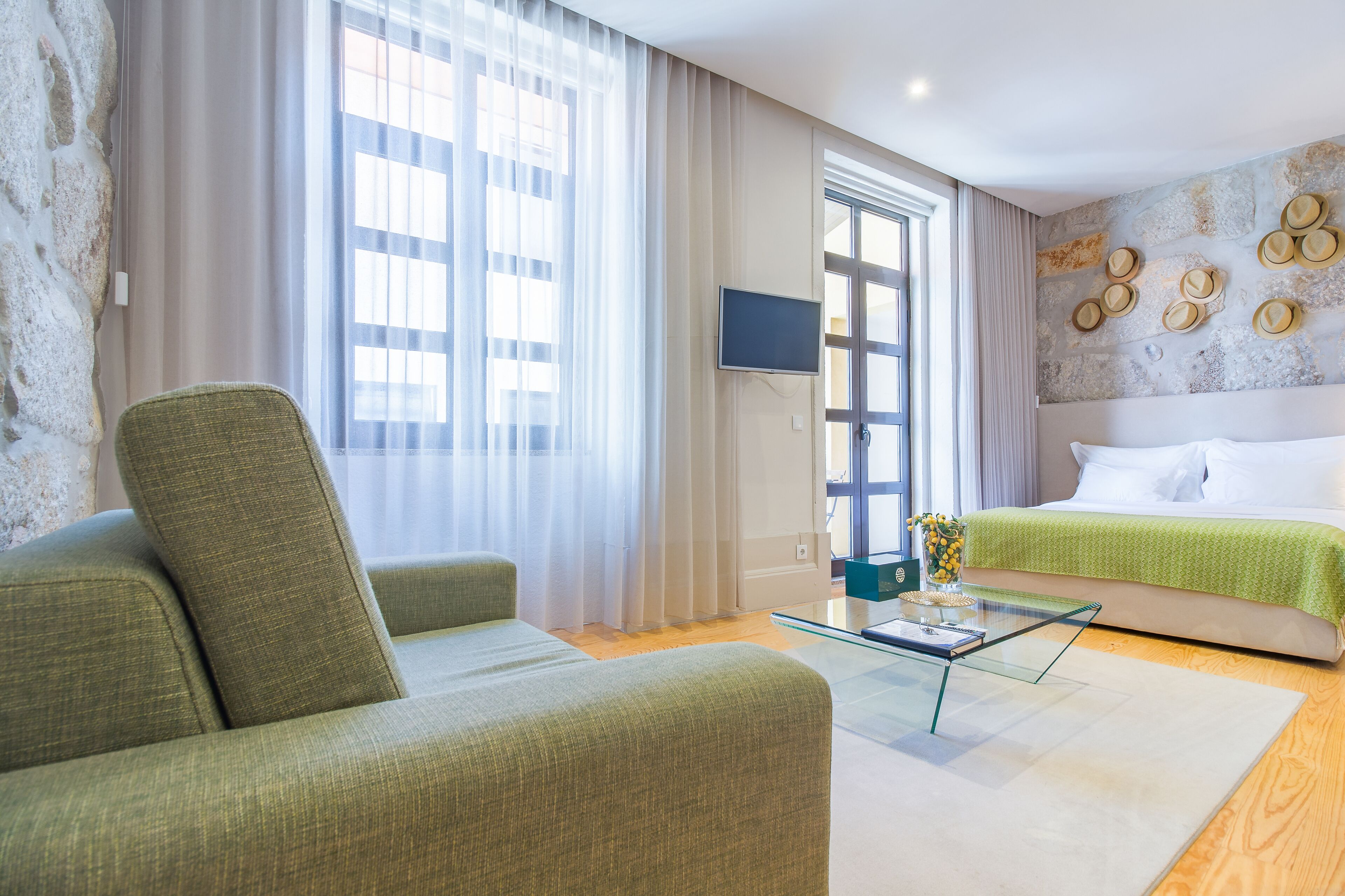 Et moderne hotelværelse med en grøn sofa, et glasbord, en seng med grønt sengetæppe, et fladskærms-TV monteret på væggen og en væg med stenudsmykning.