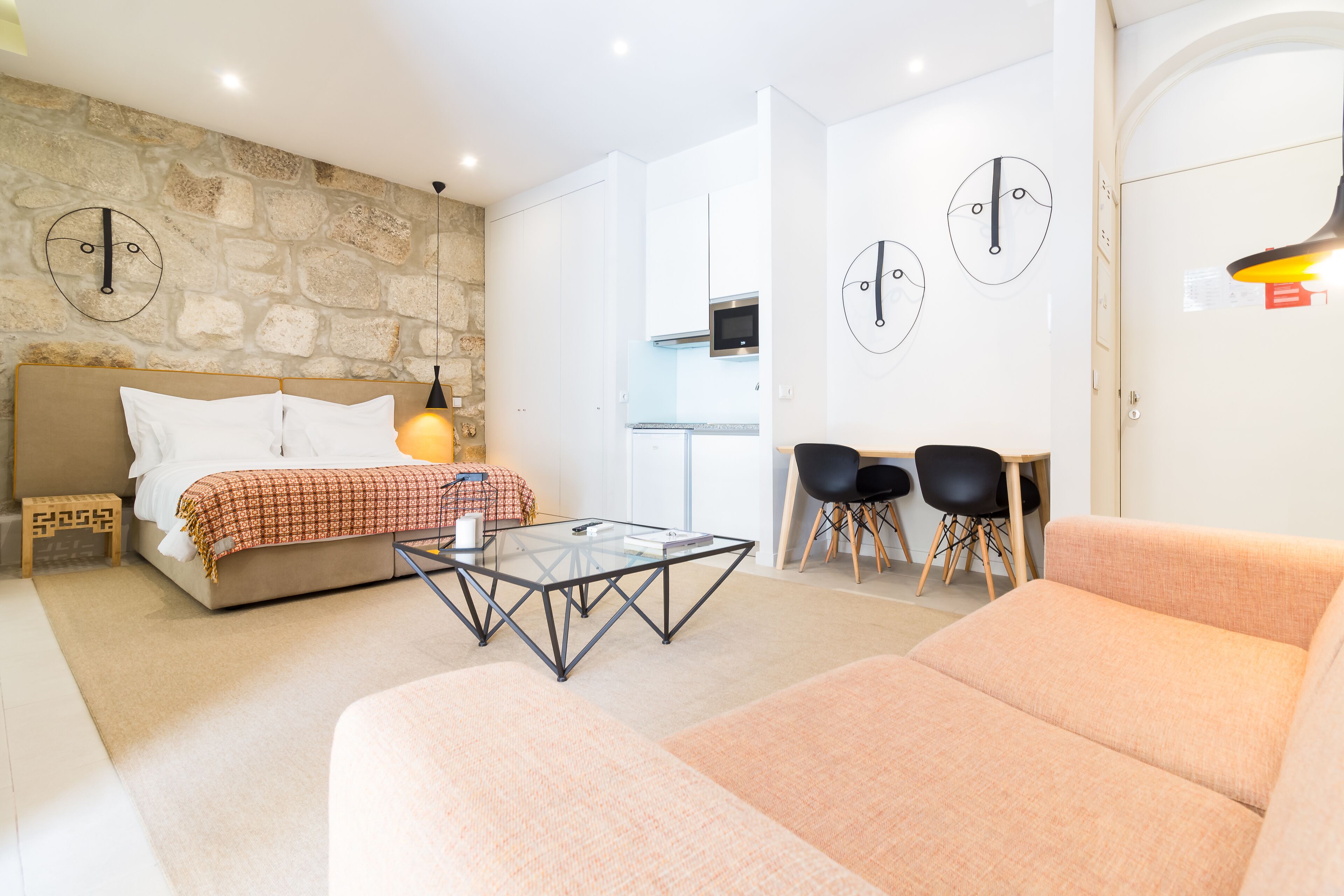 Une chambre moderne avec un mur en pierre, un lit recouvert d’une couverture à carreaux, une table basse en verre, un canapé et un bureau avec deux chaises.