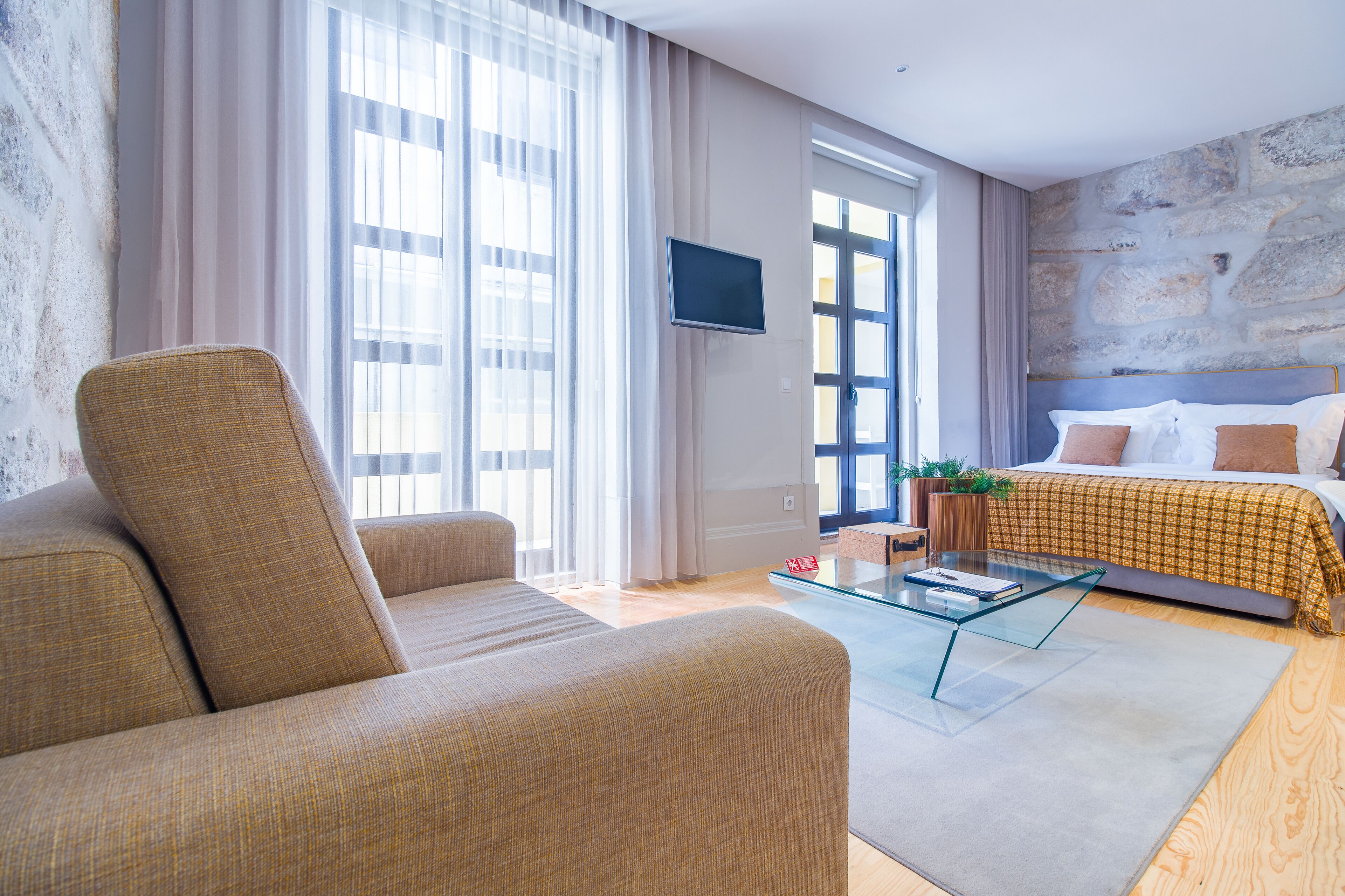 A modern hotel room with a sofa, a glass coffee table, a bed with a patterned bedspread, and a stone accent wall.