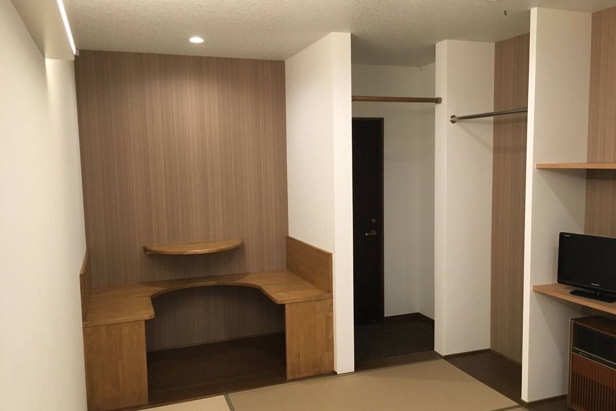 Une pièce de taille modeste, avec une coiffeuse en bois, une petite étagère, une porte donnant sur une autre pièce et un téléviseur posé sur un meuble en bois.
