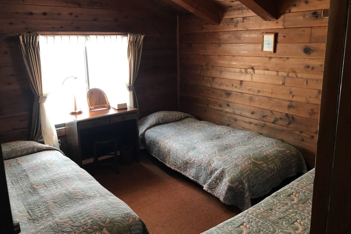 A cabin-style room with two single beds, a desk with a chair, a lamp, and a window with curtains.