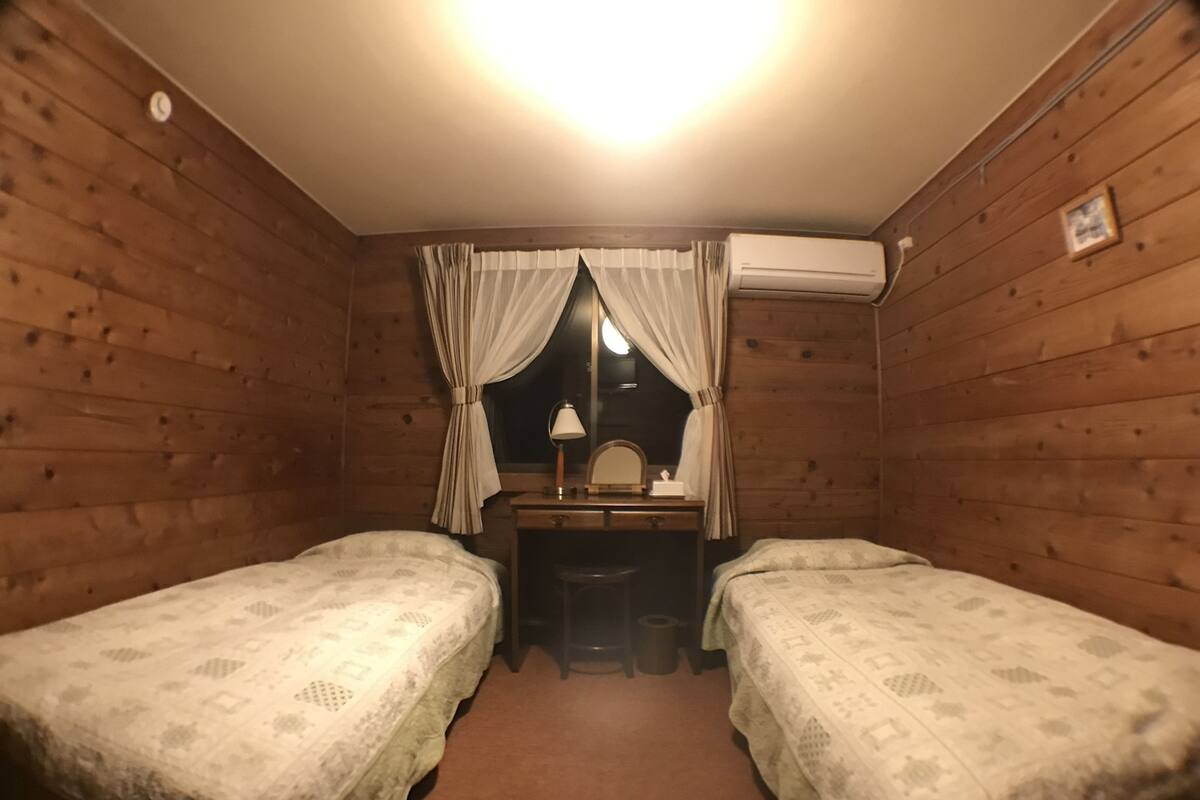 A small, cozy room with two single beds, a desk with a mirror, and an air conditioning unit on the wall.