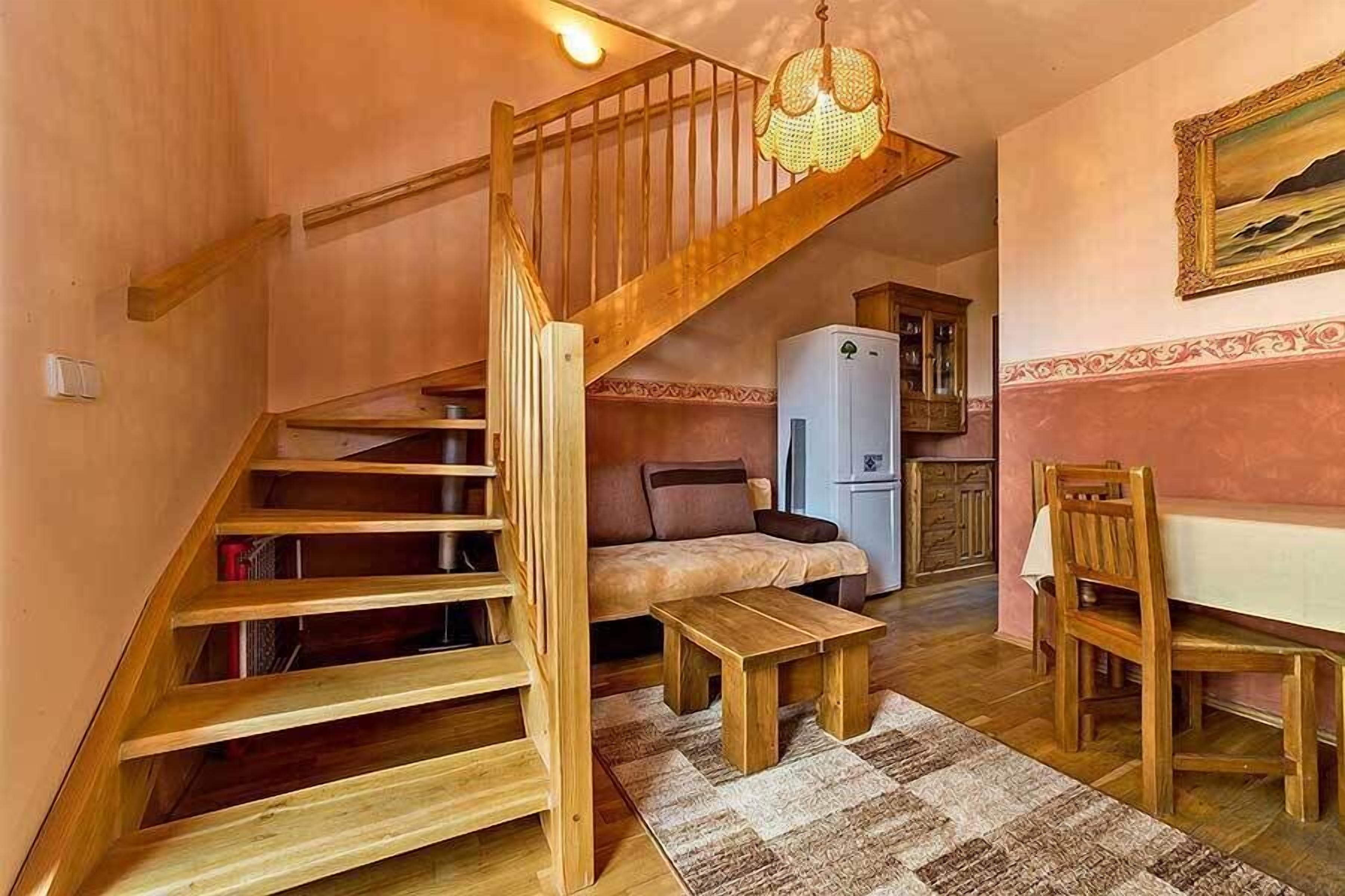 Ein kompakter Wohnraum mit einer Holztreppe, einem Sofa, einem Esstisch mit Stühlen und einem Kühlschrank.