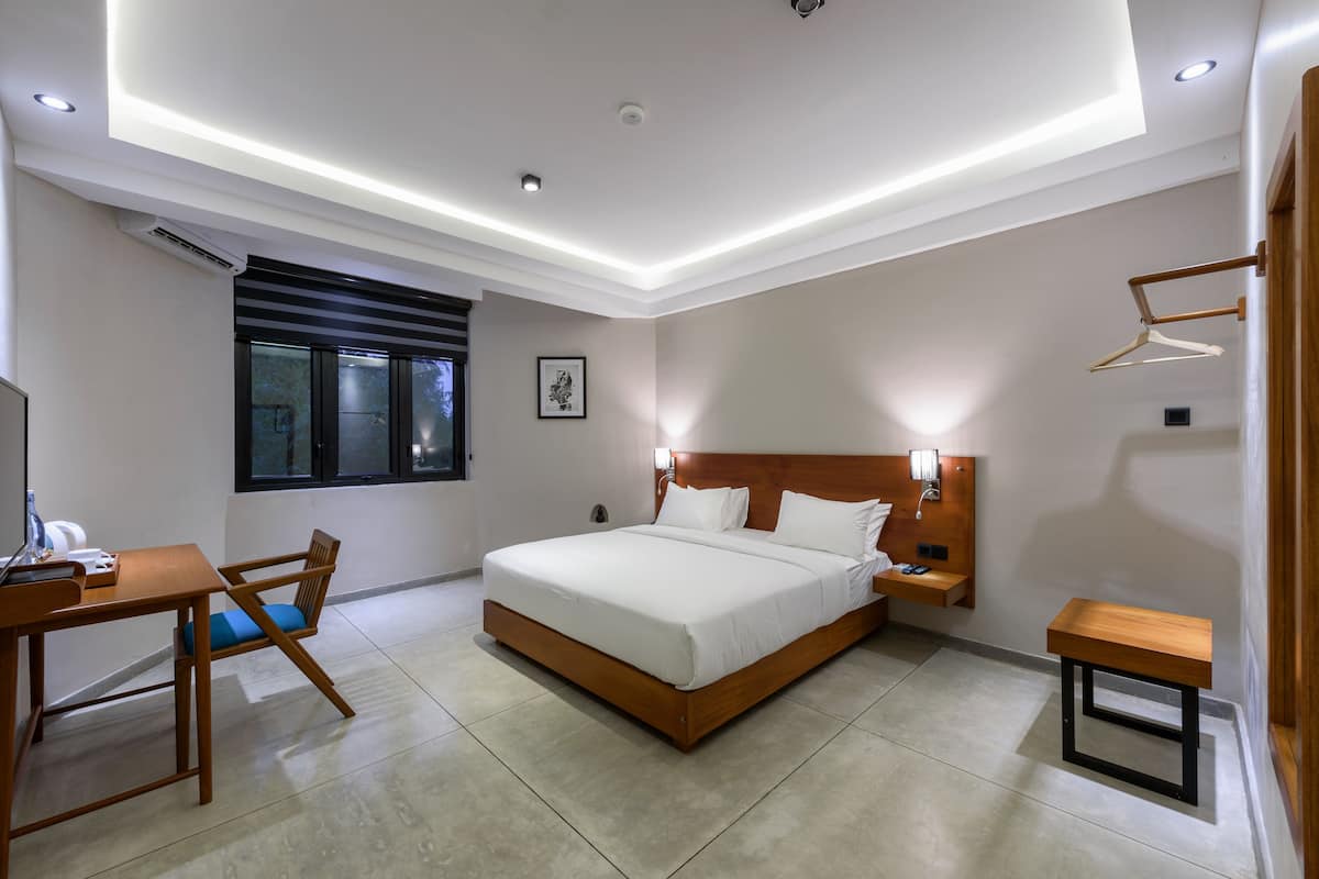 A modern hotel room with a large bed, bedside tables, a desk with a chair, and a window with blinds.