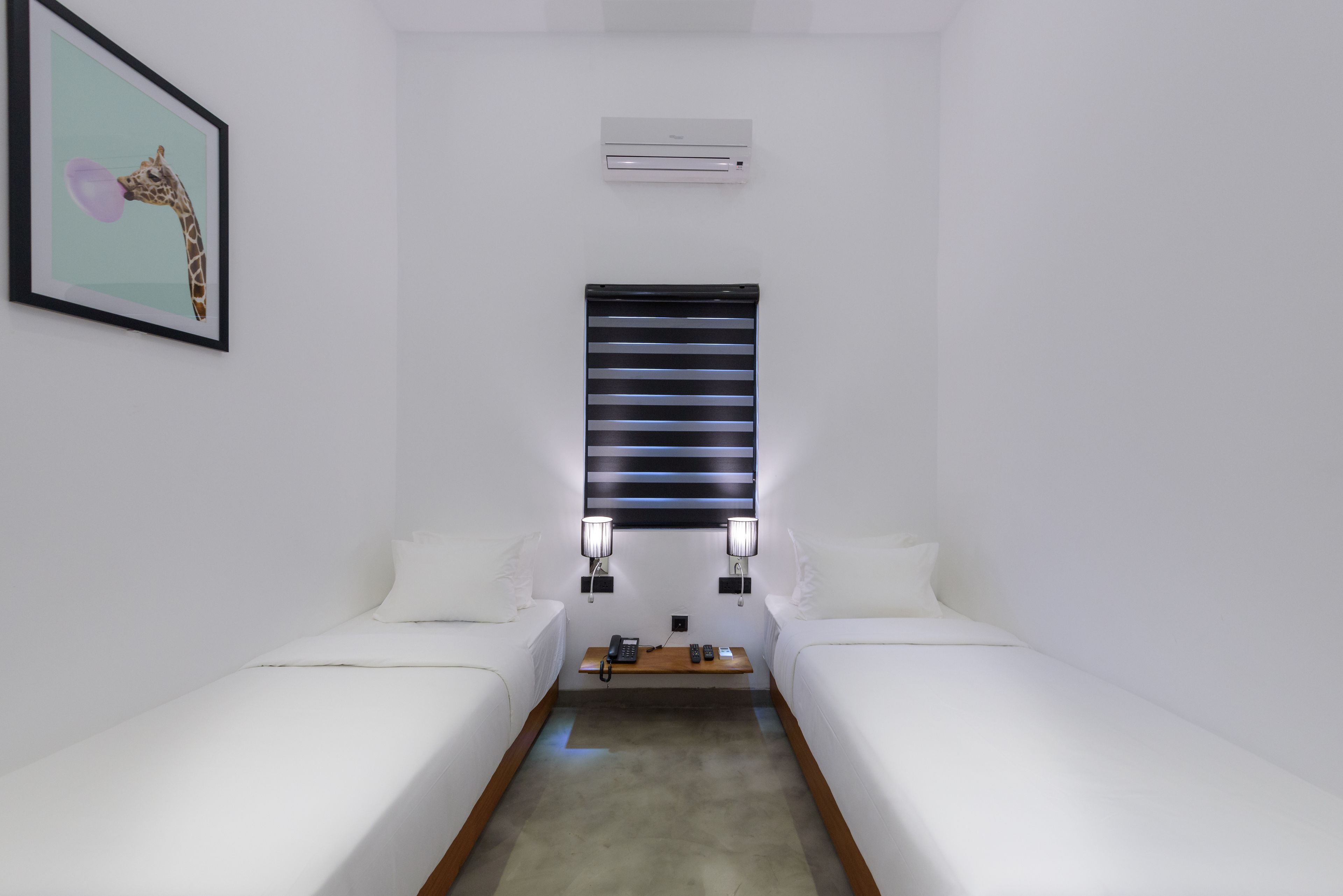 A small, clean room with two single beds, a bedside table with a phone and remote, a framed picture on the wall, and a window with blinds.